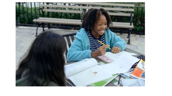 Two middle school girls studying outside with books and worksheets, collaborating and smiling during a tutoring session.