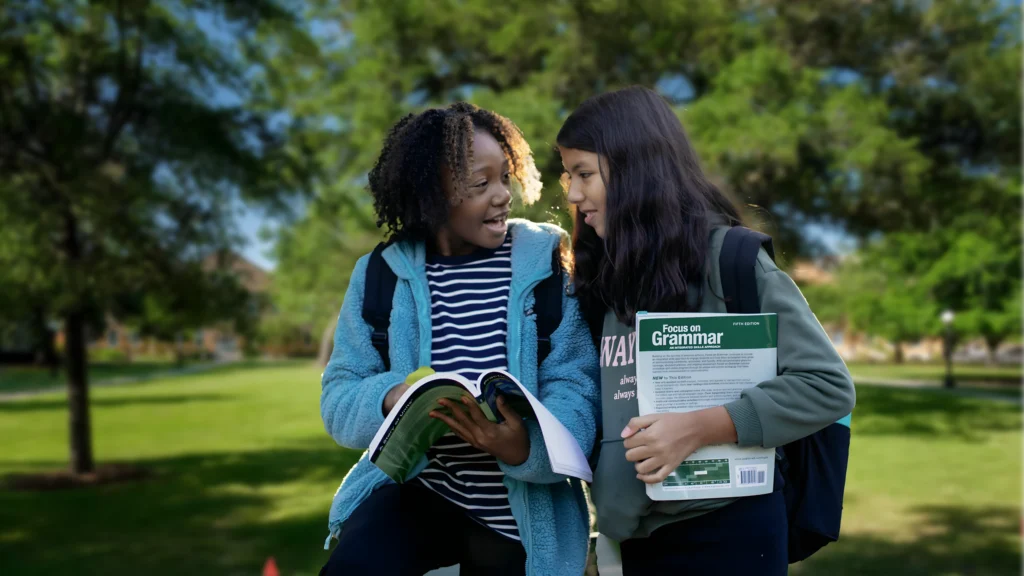 Two students with bagpacks discussing schoolwork and carrying grammar textbooks while walking through a school campus